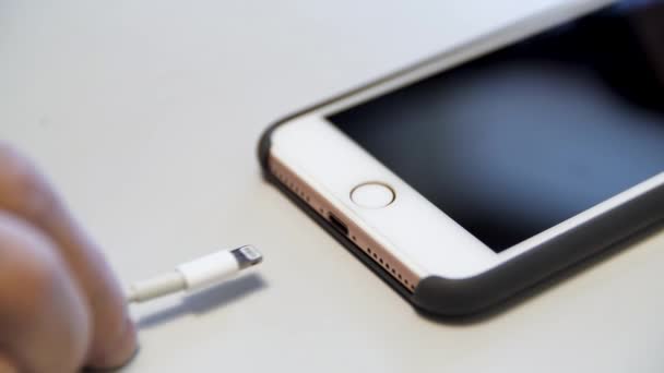 Primer plano del iphone de manzana con el cable de carga acostado en la mesa blanca. Medios. Proceso de carga de un dispositivo moderno conectado al cable USB. — Vídeos de Stock