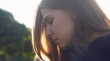 Günbatımı ve güneş ışığının arka planında genç bir kadın var. Medya. Siyah örgü kazaklı çekici bir kız kameraya bakıyor, saçları rüzgarda uçuşurken gülümsüyor..