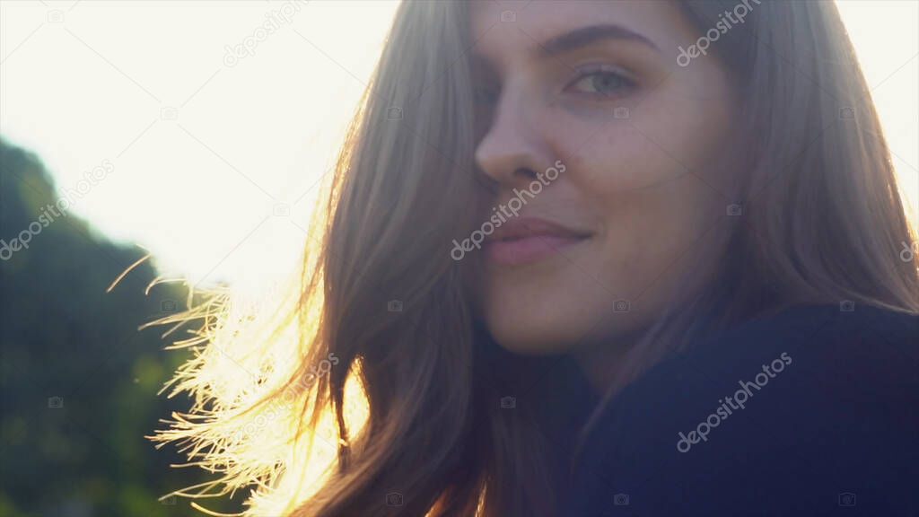 Stunning girl face against sun with her hair blowing in the wind. Media. Close up of young woman posing outdoors, smiling to the camera and touching her hair.