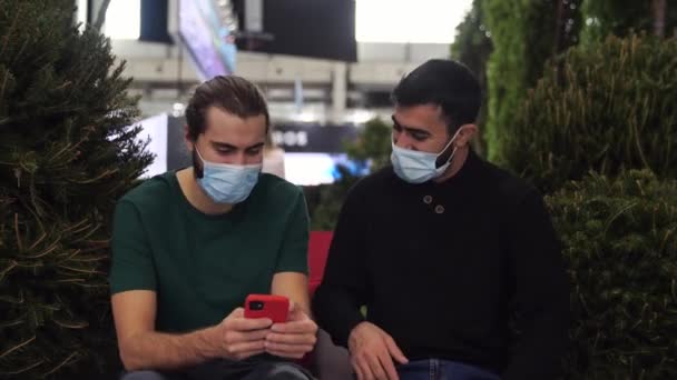 医療マスクをした男達が男性の手に装置を見て笑っている。メディア。スマートフォンでメディアコンテンツを見ている2人の友人と広告付き画面上の緑の茂みに座って — ストック動画