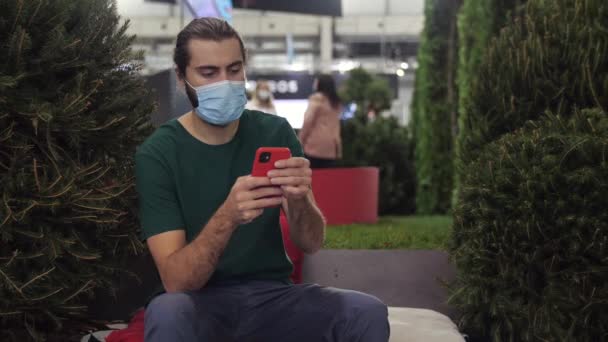 Interiér moderní výstavy. Média. Muž v zelené tričko a lékařskou masku sedí v blízkosti zelených umělých stromů a keřů a pomocí svého smartphonu. — Stock video