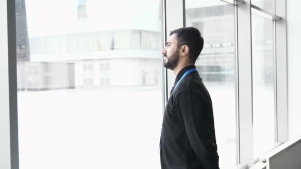 一个沉思的年轻人站在窗边的侧视图。媒体。一位挂着论坛与会者蓝色姓名标签的商人看着窗外飘落的雪花. — 图库视频影像