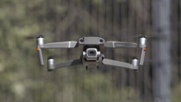 Drone with a camera hovers in the air. Action. Close up of an aircraft and blades that rotate in slow motion on blurred background of green trees, shooting process. — Stock Video