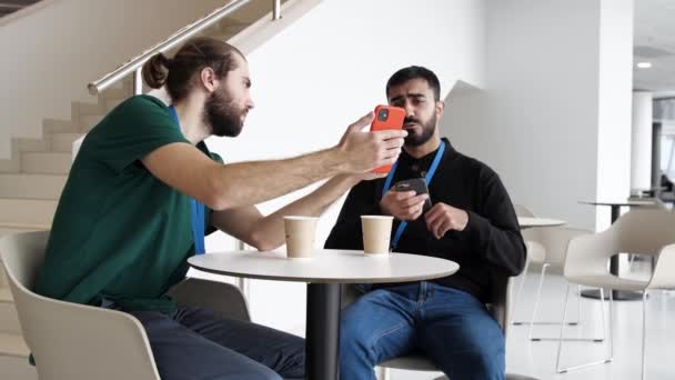 Dos jóvenes empresarios sentados a la mesa con tazas de café y discutiendo noticias. Medios. Dos hombres serios discuten el asunto de negocios en teléfonos móviles, tecnología de comunicación y concepto de negocio. — Vídeos de Stock