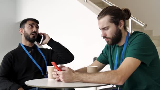 Twee jonge zakenmannen zitten aan tafel met een kop koffie en praten over nieuws. De media. Twee serieuze mannen bespreken zakelijke zaken op mobiele telefoons, communicatietechnologie en business concept. — Stockvideo