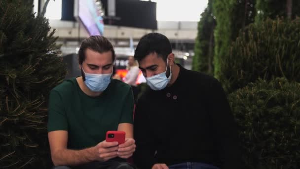 Tıbbi maskeli adamlar gülüyor ve erkeklerin elindeki alete bakıyor. Medya. İki arkadaş akıllı bir telefondan medyanın içeriğini izliyorlar ve yeşil çalıların yanında reklamlarla ekranlarda oturuyorlar. — Stok video