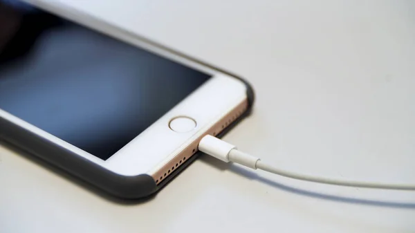 Primer plano del iphone de manzana con el cable de carga acostado en la mesa blanca. Medios. Proceso de carga de un dispositivo moderno conectado al cable USB. — Foto de Stock