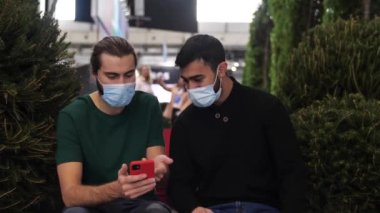 İki adam akıllı telefondan bir şeye bakıyorlar ve mutlu görünüyorlar. Medya. Tıbbi maske takan erkek arkadaşlar, arka planda teknik bir sergi ile dinleniyorlar..