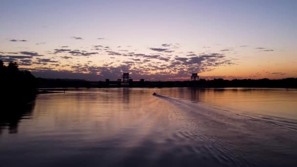 Reflexão de um pôr-do-sol laranja na água de um lago da cidade. Filmagem. Silhueta escura de árvores crescendo na costa e um barco a motor deixando rastro ondulado em um fundo de pôr do sol. — Vídeo de Stock