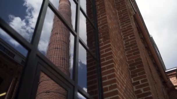 Oude schoorsteen van Moskou Arma zakenwijk weerspiegeld in een raam. Actie. Onderaanzicht van een rode bakstenen muur van een oude gasfabriek met een hoge schoorsteen op blauwe bewolkte lucht achtergrond. — Stockvideo