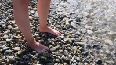 Güneşli bir yaz gününde kadın ayakları ılık deniz dalgalarıyla yıkanır. Kavram. Çakıl taşlı sahilde duran siyah tırnaklı güzel kadın ayaklarının manzarası..