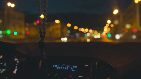 Πίνακας αυτοκινήτων στο παρασκήνιο της νυχτερινής πόλης. Στικ. Χάντρες κρέμονται στον καθρέφτη στο αυτοκίνητο σε θολή φόντο της νυχτερινής πόλης. Άποψη της πόλης νύχτα από όρθιο αυτοκίνητο — Φωτογραφία Αρχείου