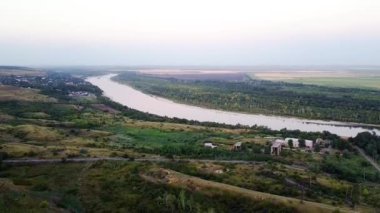 Bulutlu havada yeşil ağaçların ve tarlaların arka planında nehir ve köy manzarası. Görüntü. Yaz ufkunun arka planında bulutlu gökyüzü olan güzel bir nehir.