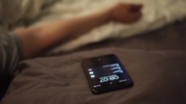 Genç adam sabahın erken saatlerinde mobil çalar saatle uyanıyor. Kavram. Uykulu adam akıllı telefondan alarmlı saat sinyalini duyduktan sonra uyanıyor, cep telefonuna uzanıyor ve onu kullanıyor..