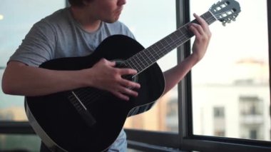 Yalnız adamın açık bir pencereye gitar çalmasına yakın çekim. Kavram. Genç müzisyen klasik akustik gitarda akorlar ve şarkılar çalıyor..