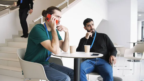 Dois jovens sérios bebendo café e conversando dentro do prédio com paredes brancas e escadas. Mídia. Parceiros de negócios confiantes discutindo ideias, planejando projetos e startups. — Fotografia de Stock