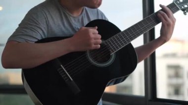 Yalnız adamın açık bir pencereye gitar çalmasına yakın çekim. Kavram. Genç müzisyen klasik akustik gitarda akorlar ve şarkılar çalıyor..