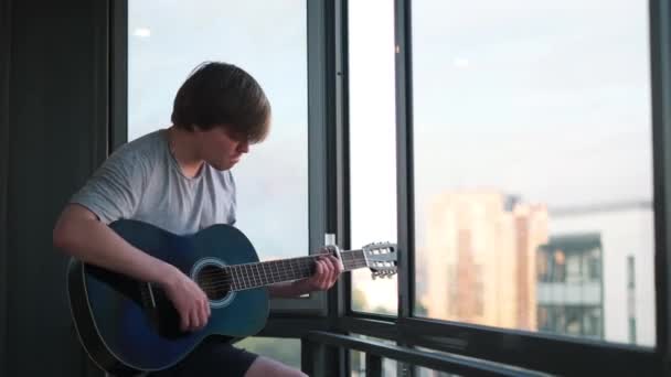 Seorang pria yang menarik berusia 20-an mengenakan kemeja abu-abu bermain gitar akustik dengan gairah. Konsep. Man bermain gitar di balkon sambil menghabiskan waktu di rumah berlatih jarak sosial. — Stok Video