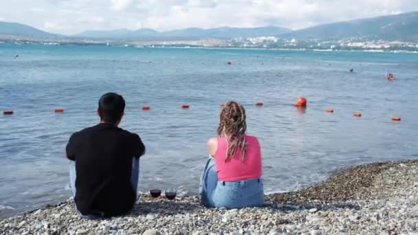 Giovane coppia con vino avendo appuntamento romantico su una spiaggia di ciottoli. Concetto. Vista posteriore di un uomo e una donna con trecce africane seduti insieme e godendo del paesaggio marino estivo. — Video Stock