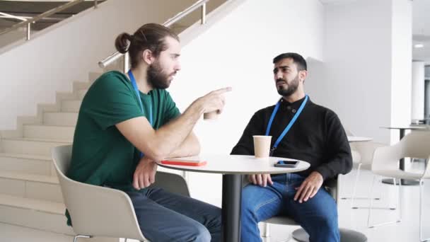 Dos jóvenes serios tomando café y hablando dentro del edificio con paredes blancas y escaleras. Medios. Socios comerciales confiados discutiendo ideas, proyectos de planificación y startups. — Vídeos de Stock