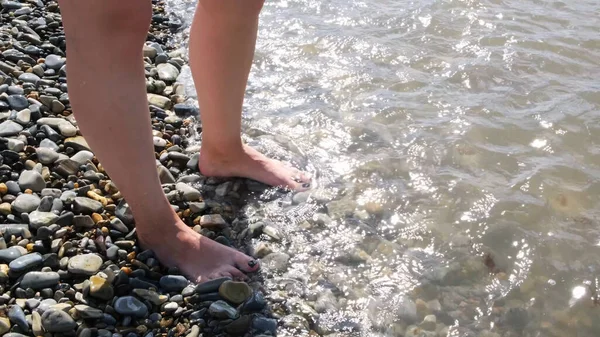 Weibliche Füße, die an einem sonnigen Sommertag von Wellen des warmen Meeres gewaschen werden. Konzept. Draufsicht auf schöne weibliche Füße mit schwarzen Nägeln, die am Kieselstrand stehen. — Stockfoto