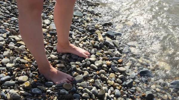 Θηλυκά πόδια πλένονται από κύματα ζεστής θάλασσας μια καλοκαιρινή ηλιόλουστη μέρα. Έννοια. Κάτοψη των όμορφων γυναικείων ποδιών με μαύρα νύχια στην παραλία με βότσαλα. — Φωτογραφία Αρχείου