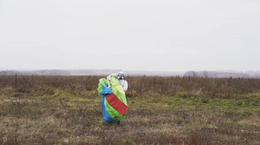 Paraşütçü sahaya indikten sonra elinde parlak bir paraşütle yürüyor. Başla. Beyaz ön cam takımlı erkek atlamacı paraşütünü taşıyor, ekstrem spor kavramı. 