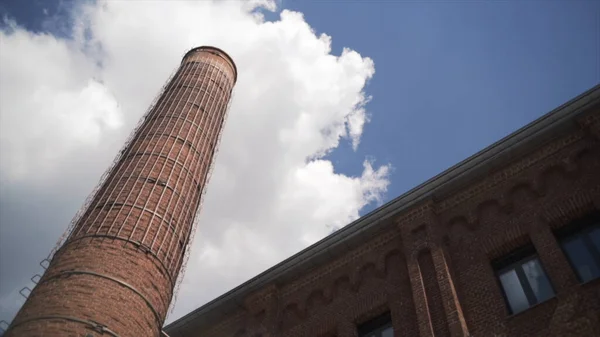 Industriële oude rode bakstenen schoorsteen op blauwe bewolkte hemel achtergrond. Actie. Onderaanzicht van een oud verlaten fabrieksgebouw met een hoge schoorsteen, concept van zware industrie. — Stockfoto