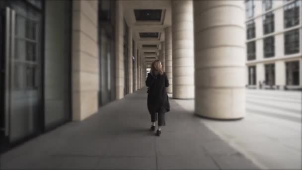 Una ragazza che camminava tra le colonne con la schiena girata verso la telecamera. Azione. Vista posteriore di una donna con i capelli ricci che cammina all'aperto vicino all'edificio con grandi finestre e pilastri giganti. — Video Stock