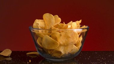 Patates cipslerinin şeffaf cam kasenin içine ve masanın üzerine düşmesini engelleyin. Stok görüntüleri. Kırmızı arka planda izole edilmiş küçük bir tabakta gevrek çöp ürünü..