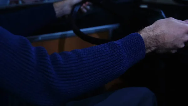 Le conducteur met la main sur un volant d'autobus. Les médias. Gros plan d'un chauffeur portant un pull tricoté bleu conduisant un bus tard dans la soirée sous la lumière d'une lanterne de rue. — Photo