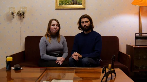 Portrét mladého sovětského páru, jak se dívá na televizi na gauči a dívá se přímo do kamery. Média. Muž a žena žijící v typickém bytě SSSR se staromódním nábytkem. — Stock fotografie