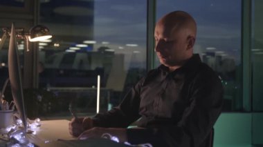 Düşünceli bir işadamı masada oturup gökdelendeki modern ofisinde yazıyor. Stok görüntüleri. Camdan duvarı ya da penceresi olan ciddi kel bir adam akşam gökyüzüne bir not yazıyor.