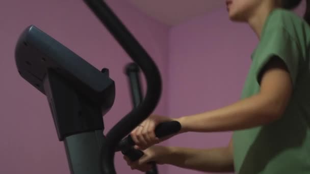 Una mujer joven está entrenando en un elipsoide, fitness y deportes en casa durante la pandemia de covidio 19. Concepto. Vista lateral de una chica haciendo deporte, estilo de vida saludable. — Vídeo de stock