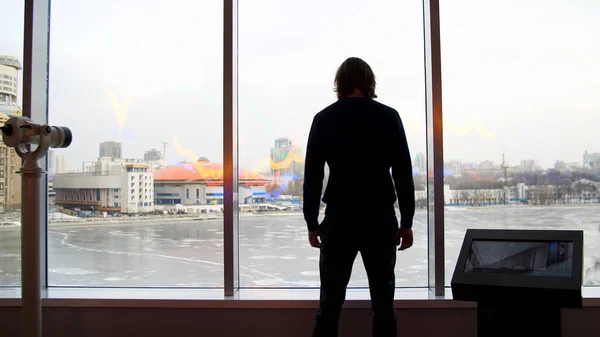 Ένας άντρας κοιτάζει μια όμορφη θέα της πόλης και μια παγωμένη λίμνη από το παρατηρητήριο. Μέσα ενημέρωσης. Οπίσθια όψη νεαρού άνδρα μέσα στο κτίριο με γυάλινους τοίχους που απολαμβάνει το χειμερινό τοπίο. — Φωτογραφία Αρχείου