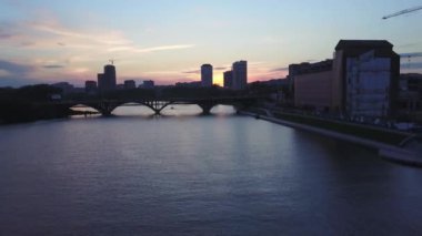 Gün batımında gökyüzü arka planında nehrin üzerinden köprüye doğru uçuyor. Stok görüntüleri. Renkli gökyüzü ve akşam şehriyle muhteşem bir manzara.