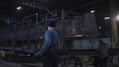 İşçi fabrikada demir yapısını elinde tutuyor. Şarjör. Adam fabrikadaki zincirlerden sallanan demir yapıyı destekliyor. Fabrika işçisi büyük bir yapı tutuyor.