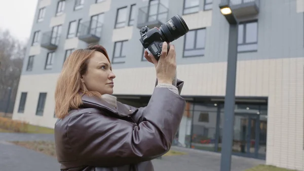 Mulher tira fotos de edifícios com câmera profissional. Acção. Mulher bonita fotógrafa dispara edifícios de vários andares. Visão traseira do fotógrafo feminino no fundo de edifícios residenciais — Fotografia de Stock