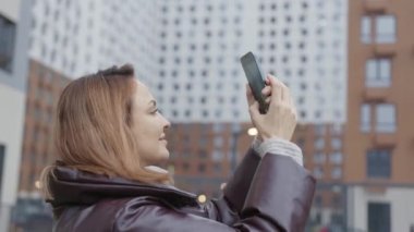 Güzel bir kadın binanın telefonunda fotoğraf çekiyor. Başla. Kadın, yüksek binaların arka planında telefonla fotoğraf çekiyor.