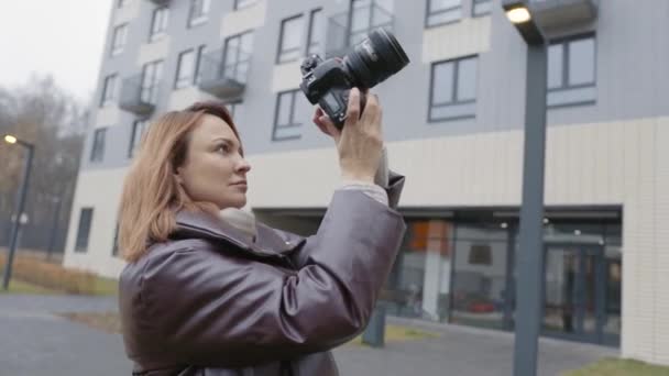 Γυναίκα παίρνει φωτογραφία σε επαγγελματική κάμερα στο παρασκήνιο του κτιρίου. Πάμε. Όμορφη γυναίκα φωτογράφος πυροβολεί αυλή του συγκροτήματος κατοικιών στην επαγγελματική κάμερα — Αρχείο Βίντεο