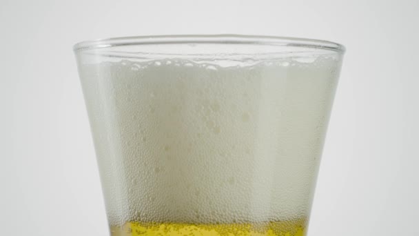 Close-up de cerveja em vidro transparente sobre fundo branco. Acção. Cerveja em vidro com espuma desaparecendo em fundo isolado. A espuma na cerveja lentamente desaparece — Vídeo de Stock
