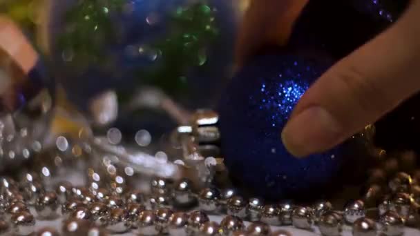 Κοντινό πλάνο του κοριτσιού χέρι βάζοντας ένα μπλε πλαστικό λάμπει χριστουγεννιάτικη μπάλα κοντά ασήμι μικρές χάντρες. Έννοια. Πρωτοχρονιάτικες διακοπές, παιχνίδια και φανταχτερή γιρλάντα για διακόσμηση ερυθρελάτης. — Αρχείο Βίντεο