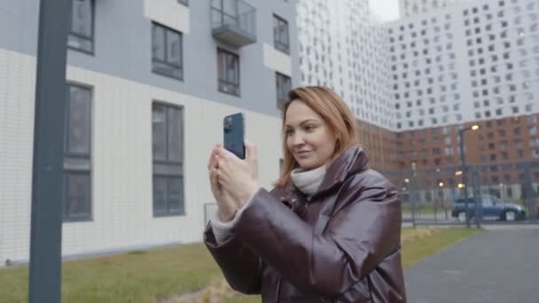 Hermosa mujer toma fotos en el teléfono del edificio. Acción. Mujer dispara y toma fotos en el teléfono en el fondo de edificios residenciales de gran altura — Vídeos de Stock