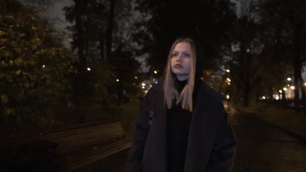 Depressed girl walking alone at night down the cold autumn street. Action. Sad blond teen woman in a coat walking along bushes and street lamps late in the evening. — Stock Video