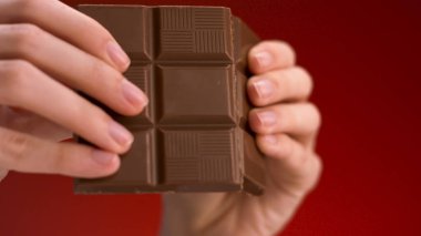 Kırmızı arka planda izole edilmiş bir çikolatayı kıran kadın ellerine yakın çekim. Stok görüntüleri. Şeker ve şekerleme kavramı. 
