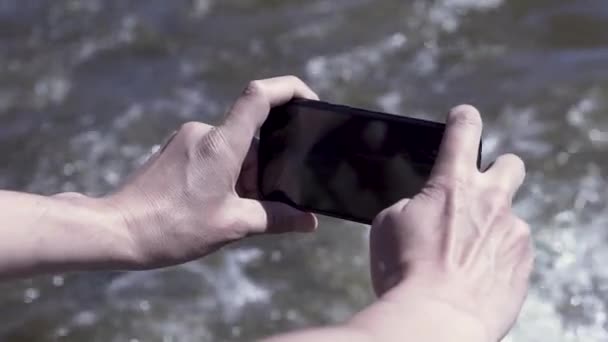 Jeune homme tenant un smartphone et enregistrant une vidéo avec un beau ruisseau fluvial. Clip. Gros plan des mains masculines prenant des photos de la rivière rapide. — Video