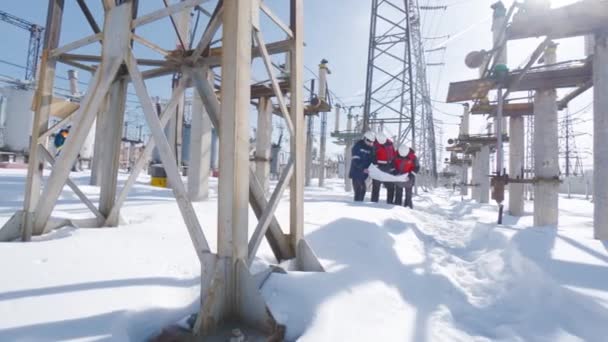 Μηχανικοί σε ηλεκτρικό υποσταθμό το χειμώνα. Πάμε. Μηχανικοί και ηλεκτρολόγοι εξετάζουν το σχέδιο ηλεκτρικών υποσταθμών το χειμώνα. Εργασία ηλεκτρικό υποσταθμό για σαφή ημέρα του χειμώνα — Αρχείο Βίντεο