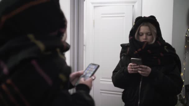 Máma používá svůj smartphone před zrcadlem, připravená vyrazit, oblečená v teplých zimních šatech. Akce. Blondýny mladá dívka textování zprávu na chodbě doma. — Stock video