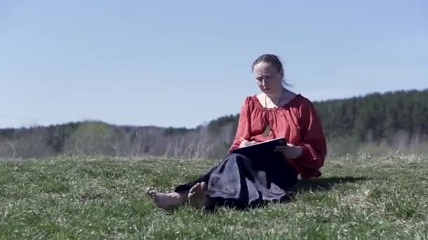 Жінка, одягнена в традиційну російську сорочку і довгий халат, сидить на луговій траві наодинці і малює. Кліп. Жінка середнього віку сидить на траві в парку променів сонця, насолоджуючись малюнком — стокове відео