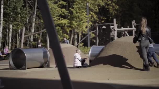 Ao ar livre parque infantil moderno com diferentes equipamentos para diversão e para atividades esportivas. Acção. Uma menina está balançando e uma mãe com seu filho estão tentando conquistar a colina artificial. — Vídeo de Stock